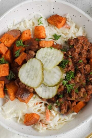 Shallow bowl with sloppy Joe mix, roasted sweet potatoes, coleslaw, and pickles with 2 gold forks on the side.