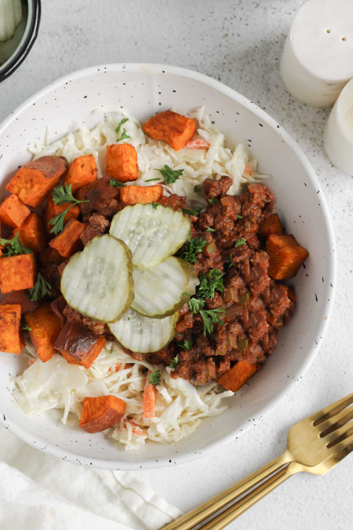 Shallow bowl with sloppy Joe mix, roasted sweet potatoes, coleslaw, and pickles with a gold fork and salt and pepper shakers off to the side.