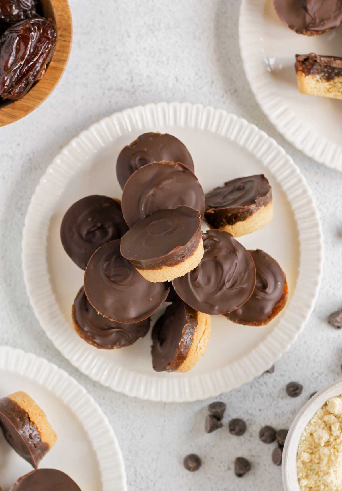 Round Twix bites stacked on a small plate with some of them laying on the side. Additional plates, a bowl of almond flour, and a bowl of dates off to the side.
